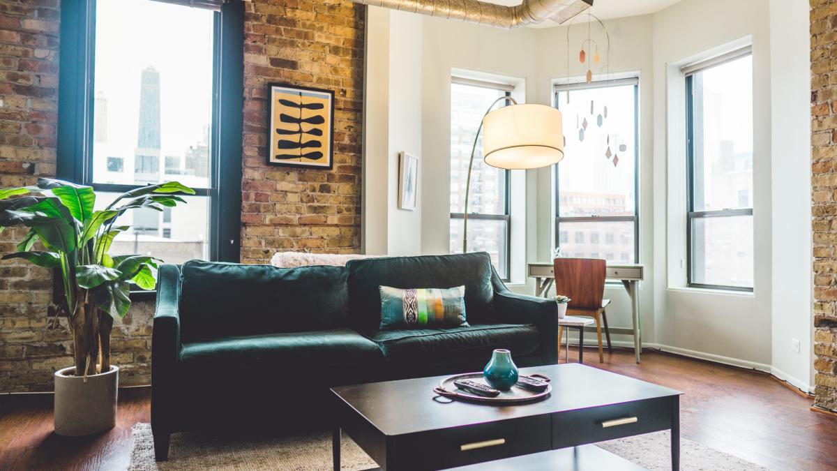 Modern loft-style living room with exposed brick walls, large windows, a dark green sofa, coffee table, indoor plant, and city views.