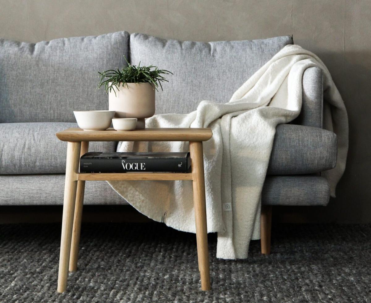 Cozy living room scene with a grey fabric sofa, wooden side table holding a potted plant and bowls, a folded throw blanket, and a book shelf underneath for a minimalist, Scandinavian look.
