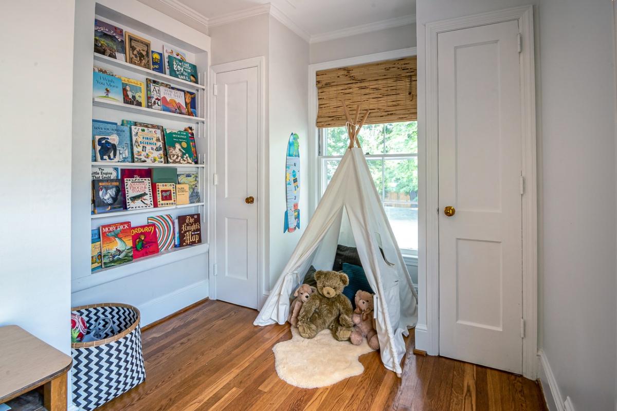 Children’s reading nook with a white teepee tent, plush toys, built-in bookshelf filled with books, and wooden flooring beside a bright window.