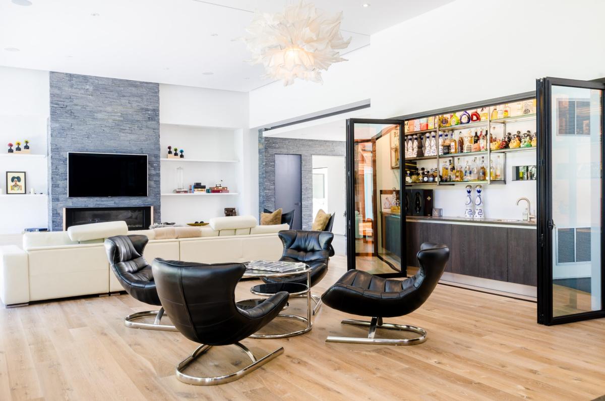 Open-plan modern living room with leather lounge chairs, neutral sofas, a wall-mounted TV, and a sleek glass-enclosed home bar with shelves of spirits.