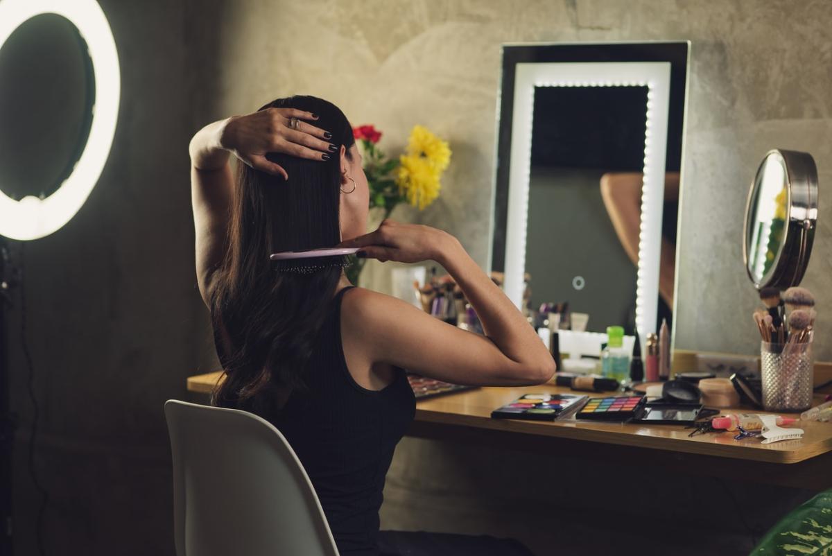 Woman brushing her hair at a vanity table with illuminated mirror, surrounded by makeup products and styling tools in a softly lit room.