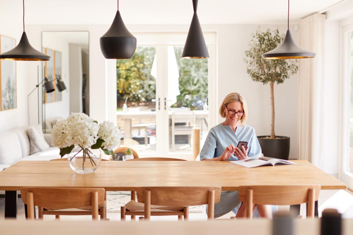 Woman sitting at a wooden dining table in a bright, modern dining room, using a smartphone beside an open book and vase of flowers.