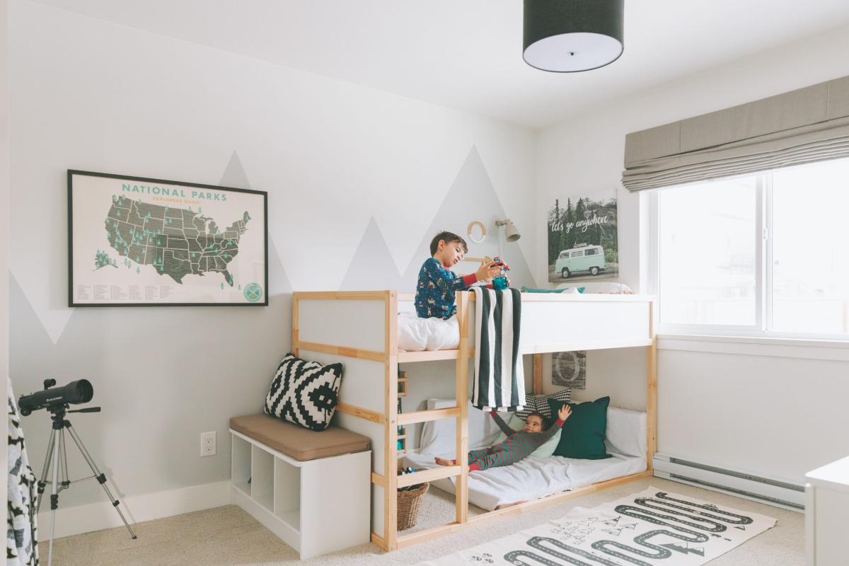 Children’s bedroom with wooden bunk beds, kids playing on the top and bottom bunks, wall art, and soft neutral décor by a bright window.
