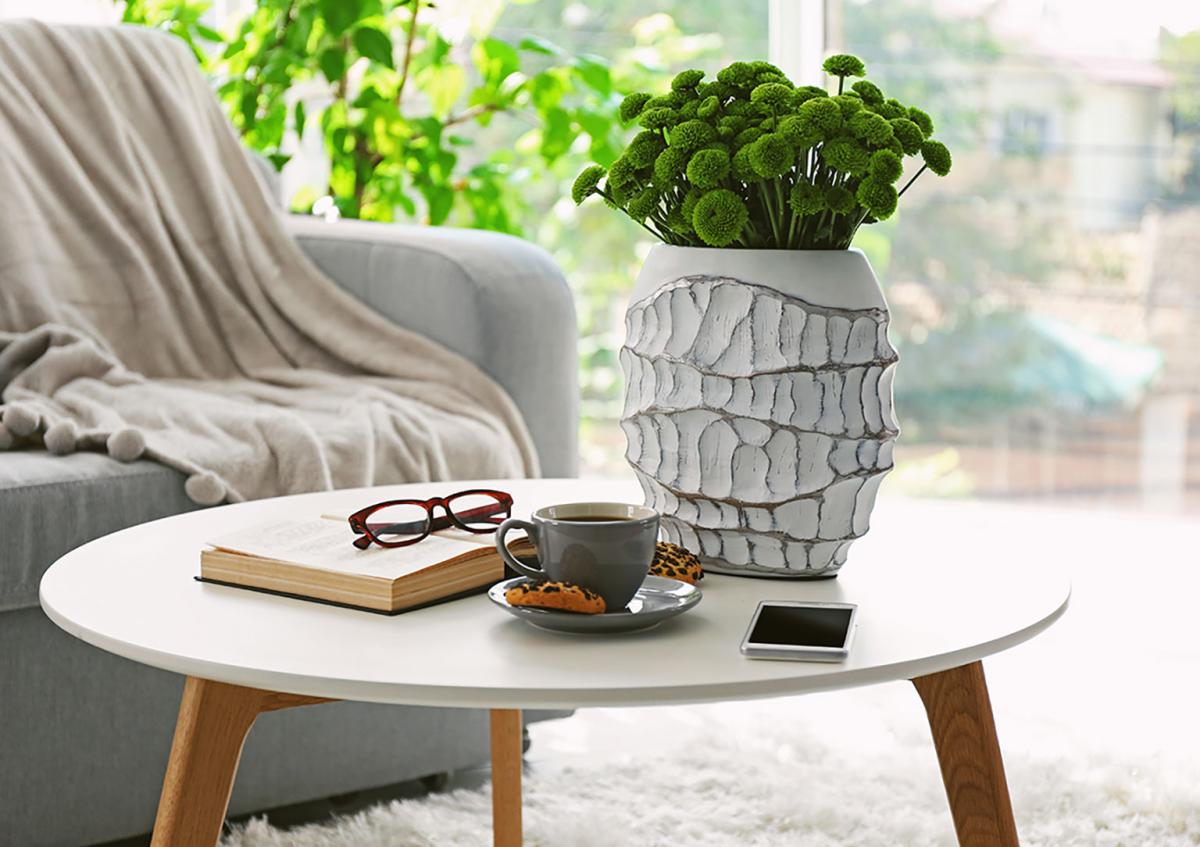 White round coffee table with a vase of green flowers, a cup of coffee, book, glasses, and smartphone in a bright living room.