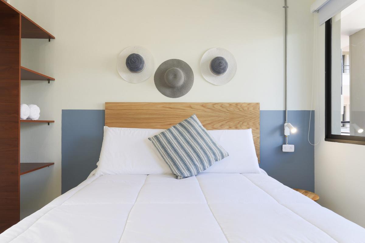 Minimalist bedroom with a wooden headboard, white bedding, blue accent wall, and decorative woven wall art above the bed, styled with modern lighting and open shelving.