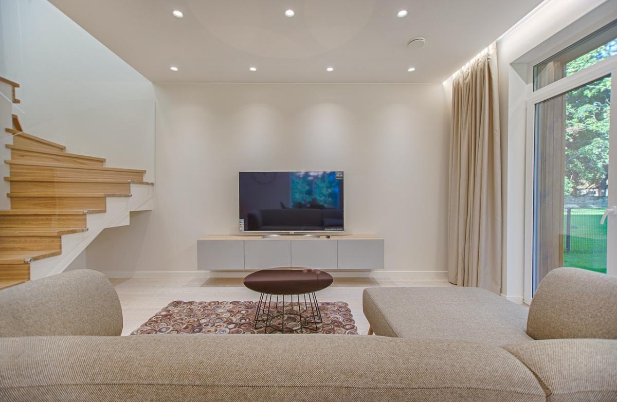 Modern living room with neutral sofa, wall-mounted TV on a low unit, wooden staircase, and large window overlooking a garden.