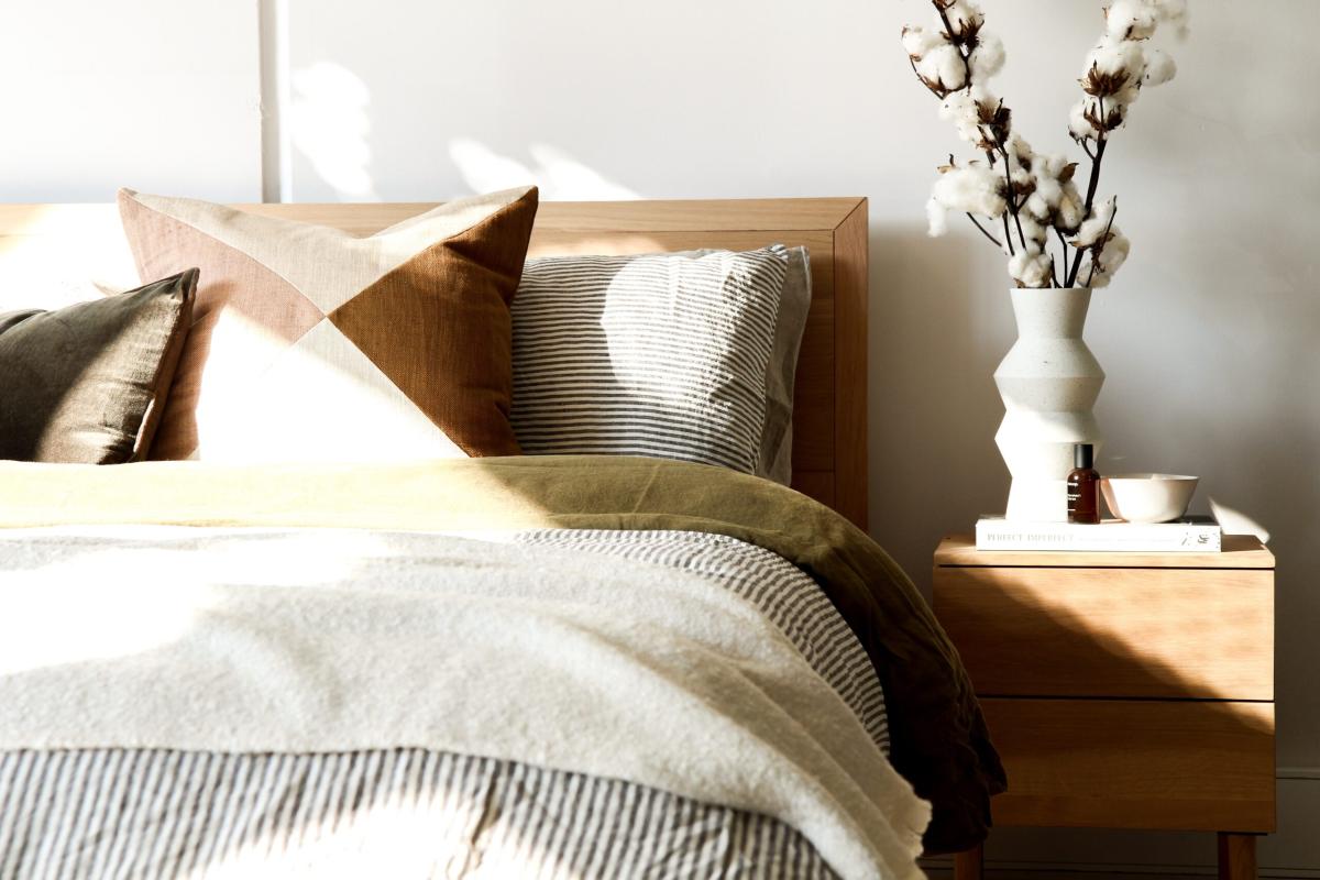 Sunlit bedroom with a wooden bed frame, layered neutral bedding, patterned cushions, and a bedside table styled with a vase and cotton stems.