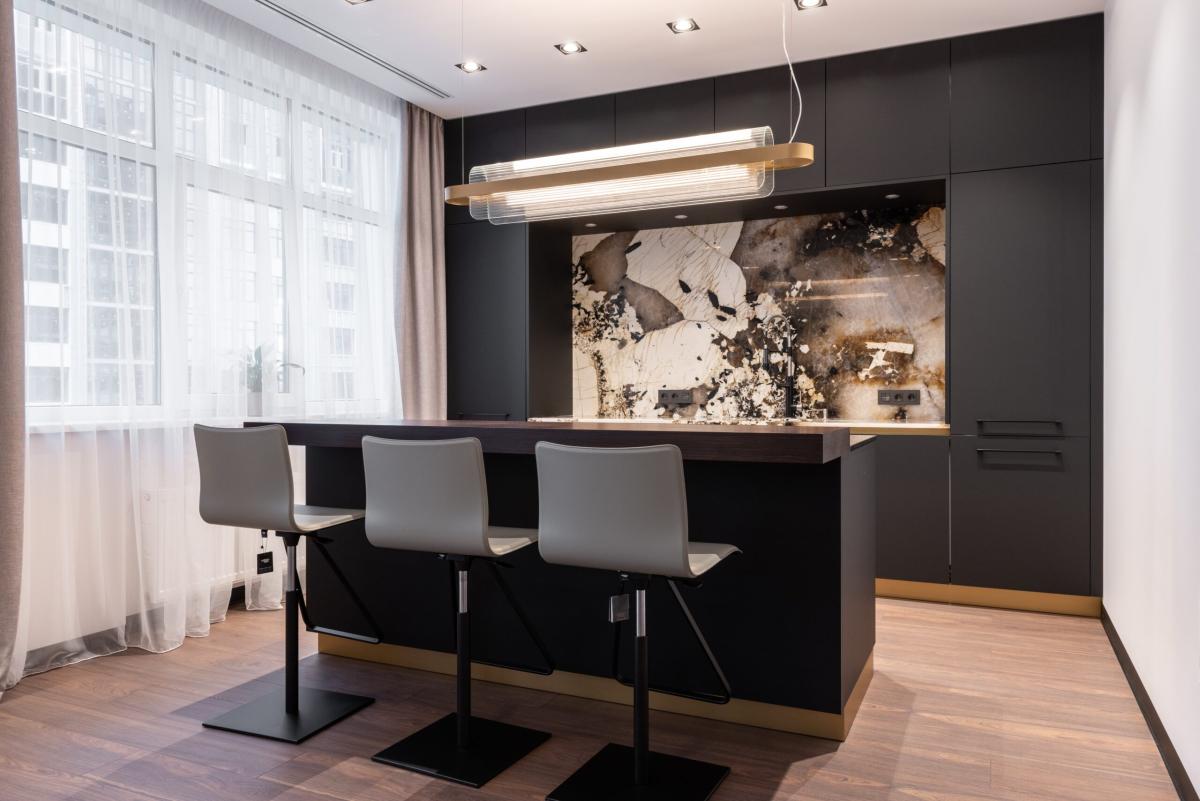 Modern kitchen with a black island, three bar stools, sleek cabinetry, pendant lighting, and a marble-effect backsplash in a contemporary apartment.