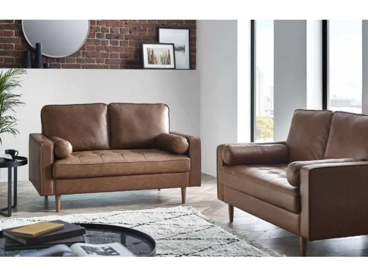Brown leather sofa and matching armchair with wooden legs, styled in a modern living room with exposed brick, neutral décor, and natural light.
