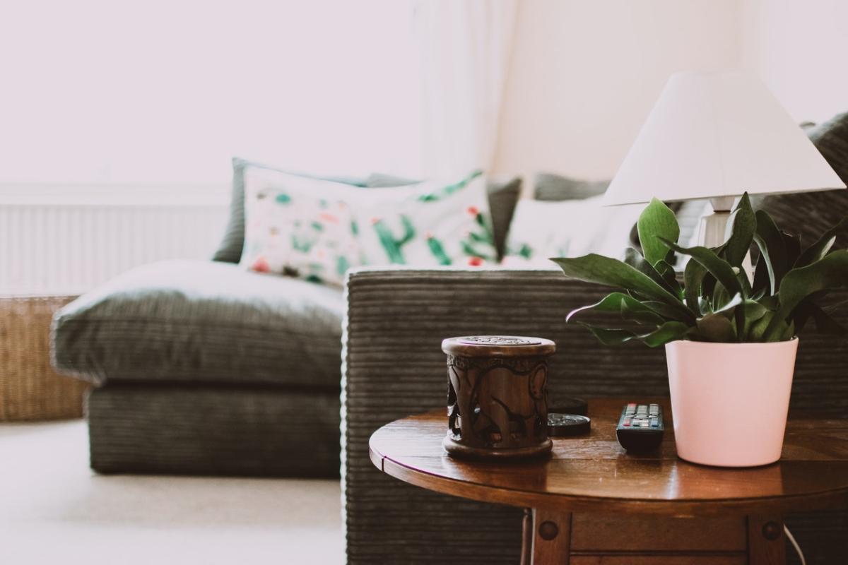 Cozy living room detail with a wooden side table holding a potted plant, candle, and remote, set in front of a grey sofa with patterned cushions.