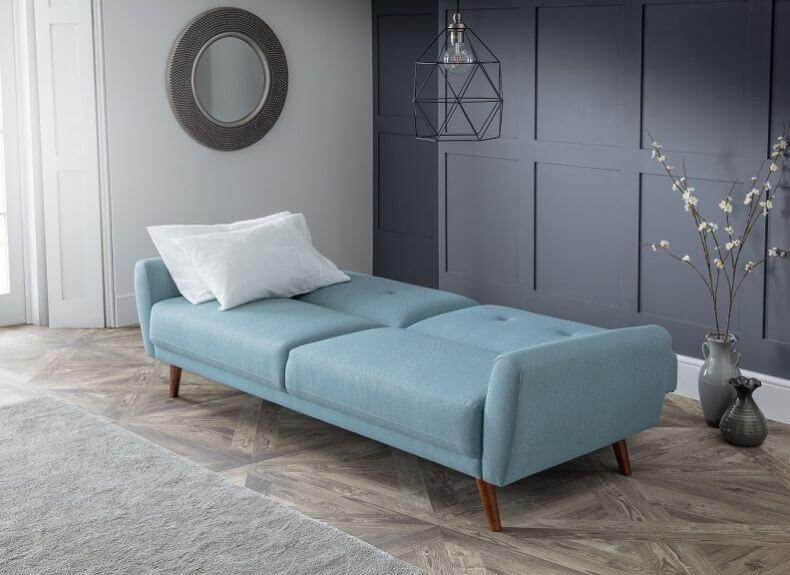 Modern upholstered chaise lounge in soft blue fabric with wooden legs, styled in a minimalist room with paneled dark walls and decorative lighting.
